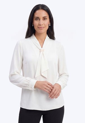 Vrouw met lang donker haar draagt een witte blouse met een parelbroche op de das en bijpassende pareloorbellen, staand tegen een effen achtergrond.