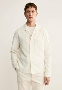 Camisa de botones color crema hecha de tela suave, con un bolsillo parche, cuello clásico y mangas largas, combinada con pantalones de un color claro a juego.