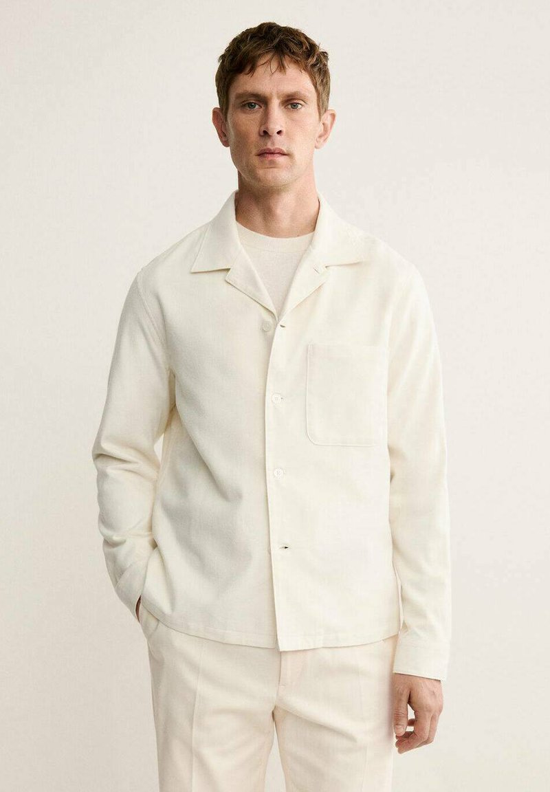 Camisa de botones color crema hecha de tela suave, con un bolsillo parche, cuello clásico y mangas largas, combinada con pantalones de un color claro a juego.