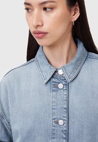 Šviesiai mėlyna džinsinė striukė su smailiu apykakliu, užsegama metalinėmis spaudėmis ir matomomis siūlėmis palei siūles.