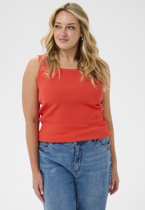 Femme blonde portant des boucles d’oreilles créoles en or, un haut rouge sans manches et un jean bleu, regardant vers sa gauche, souriant légèrement.