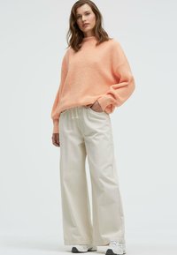 Light coral knit sweater with a high neckline, paired with cream wide-leg trousers and white sneakers. Minimalist and relaxed fit.