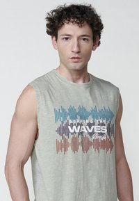 Gaiši zaļa bezpiedurkņu krekls, kas izgatavots no mīksta auduma, ar grafiku, kurā attēloti krāsaini skaņas viļņu modeļi un teksts "SPLAVOJOT VIĻŅUS."