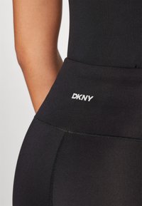 Svart leggings laget av tøynbart materiale, med høy midje og en sydd søm, samt en hvit "DKNY" logo på siden.