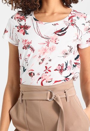 Femme portant un haut blanc à manches courtes avec un imprimé floral rose et rouge rentré dans un pantalon beige taille haute avec une ceinture nouée.