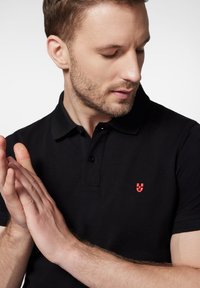 Czarna koszulka polo z kołnierzykiem i krótkimi rękawami, z małym czerwonym logo po lewej stronie. Gładka tekstura materiału.