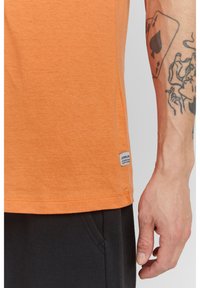 Rövid ujjú, narancssárga pamut ing egyenes szabással, kis címkével a szegélyen és sima textúrával. Fekete melegítőnadrággal viselve.