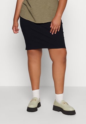 Kobieta ubrana w czarną spódnicę do kolan, oliwkowozielony top, białe skarpetki i beżowe platformowe mokasyny stoi na tle jednolitego tła.