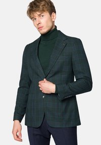 Zielona marynarka w kratę o dopasowanym kroju, z pojedynczym zapięciem na guzik, klapami wyprofilowanymi i textured tkaniną. Noszona na ciemnym golfie.