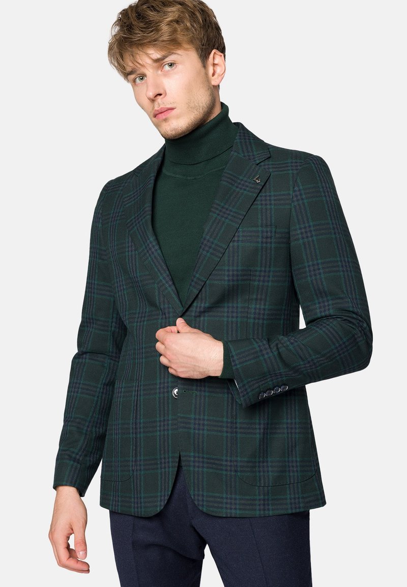 Zielona marynarka w kratę o dopasowanym kroju, z pojedynczym zapięciem na guzik, klapami wyprofilowanymi i textured tkaniną. Noszona na ciemnym golfie.
