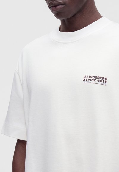 Άνδρας που φοράει λευκό T-shirt με κοντά μανίκια και λογότυπο "J. LINDEBERG ALPINE GOLF" στο αριστερό μέρος του στήθους.