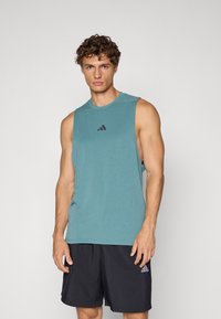 Teal ærmeløs træningsskjorte lavet af blødt stof, med en rund halsudskæring og et sort Adidas-logo på brystet. Sammen med sorte shorts.