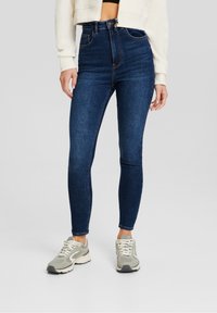 Jeans skinny de tiro alto en azul oscuro con una textura suave, bolsillos delanteros y costuras sutiles. Combinados con zapatillas texturizadas en gris claro.