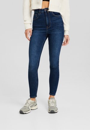 Jeans skinny de tiro alto en azul oscuro con una textura suave, bolsillos delanteros y costuras sutiles. Combinados con zapatillas texturizadas en gris claro.