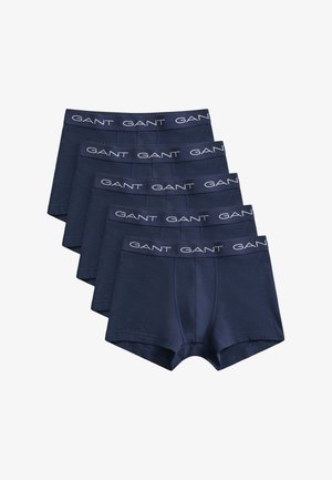 Muž bez košele s kučeravými tmavými vlasmi, oblečený v námornícky modrých boxerkách s pásom s logom „GANT“, stojaci na jednoduchom svetlom pozadí.