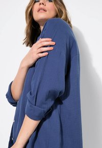 Camicia blu con texture, dettagli sui bottoni e maniche arrotolate. Il tessuto appare morbido e leggermente stropicciato, offrendo una vestibilità rilassata.