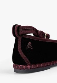 Zapato de terciopelo negro con acentos burdeos, que presenta un logotipo decorativo de calavera, un ribete trenzado y una puntera redondeada. Diseño minimalista.