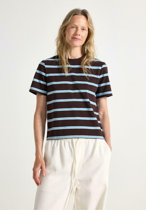 Kvinde med langt blondt hår iført en brun T-shirt med lyseblå og hvide vandrette striber og løstsiddende hvide bukser med snører.