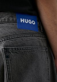 Grå denimjeans har en blå etikett med "HUGO" i vit text på baksidan. Tyget har en texturerad look med synlig sömnad.