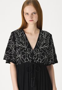Rochie neagră cu decolteu adânc în V, cu modele florale brodate în alb pe mâneci și o talie cu fundiță și ciucuri. Tesătură texturată.
