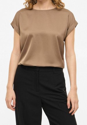 Frau trägt eine kurzärmelige Satinkleid-Bluse in Taupe, die in hoch taillierte schwarze Hose gesteckt ist, vor einem schlichten Hintergrund.