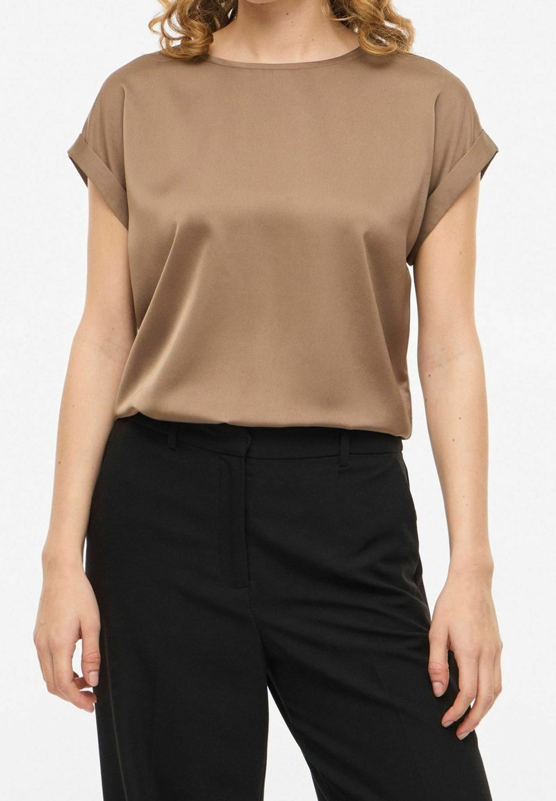 Femme portant un chemisier en satin taupe à manches courtes rentré dans un pantalon noir taille haute sur un fond uni.
