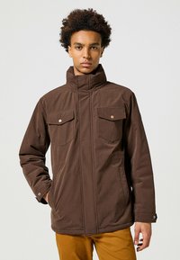 Giacca isolante marrone con colletto alto, due tasche sul petto e una trama di tessuto resistente. Caratterizzata da bottoni a pressione e un design comodo.