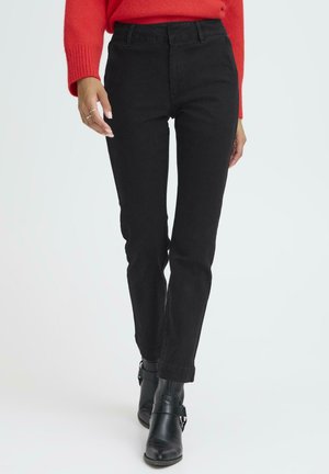 Frau in einer engen schwarzen Hose, schwarzen Stiefeletten und einem roten Pullover, die vor einem schlichten hellen Hintergrund geht.