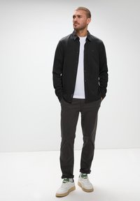 Svart knappad skjorta över en vit t-shirt, ihopparad med mörkgrå byxor och vita skor med gröna accenter. Slät textur, avslappnad passform.