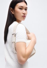 Νεαρή γυναίκα με λευκό κοντομάνικο τοπ με υφή, κρατώντας μία μπεζ τσάντα στον ώμο της, κοιτάζοντας πλάγια με ουδέτερη έκφραση.