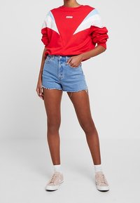 Červená mikina s modrými a bílými akcenty, doplněná džínovými šortkami. Modelka má na sobě růžové tenisky a bílé kotníkové ponožky. Ležérní outfit.