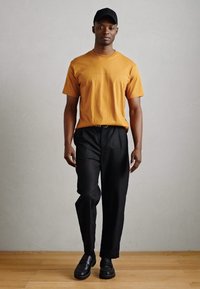 Oranssi puuvillapaita, mustat housut ja mustat kengät. Henkilöllä on päässään musta lakki. Sileä pinta, yksinkertainen muotoilu, neutraali tausta.