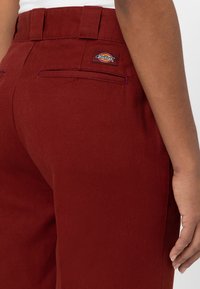 Burgunderfärgade byxor i slitstark tyg, med bakfickor som har en märkeslogotyp och kontrasterande söm längs sidan.