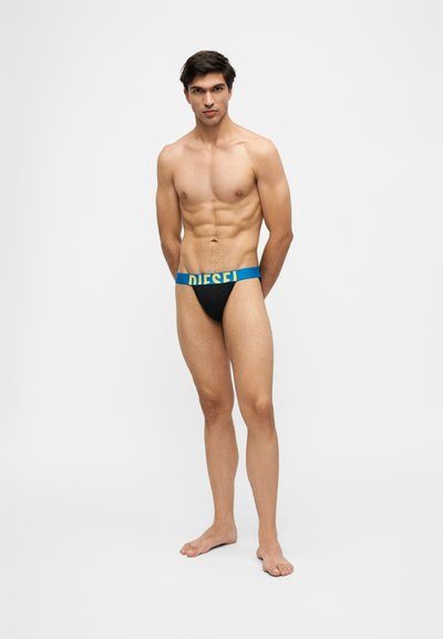 Homme musclé debout, pieds nus, portant un slip noir avec une large ceinture bleue portant l'inscription "DIESEL" sur un fond clair uni.