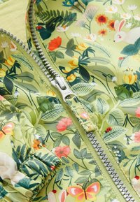 Veste zippée florale en vert clair, arborant des motifs botaniques de différentes couleurs, un zip en argent et des poignets froncés. Texture de tissu lisse.