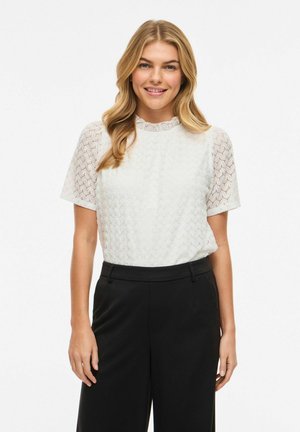 Femme aux longs cheveux blonds portant un chemisier blanc en dentelle à manches courtes et un pantalon noir taille haute, souriant à la caméra.