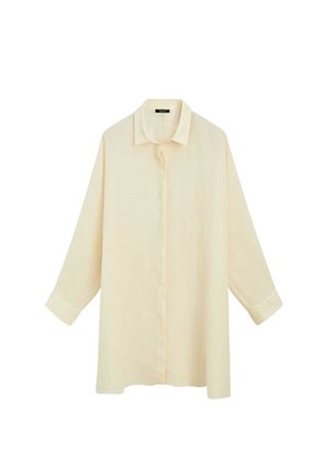 Femme portant une chemise beige clair surdimensionnée, un pantalon à jambes larges, des lunettes de soleil et un long collier, tenant un sac tressé sur un fond uni.