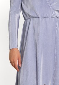 Main d’une femme reposant sur le côté, portant une robe brillante, côtelée, mauve clair à manches longues avec une taille cintrée et un ourlet asymétrique.