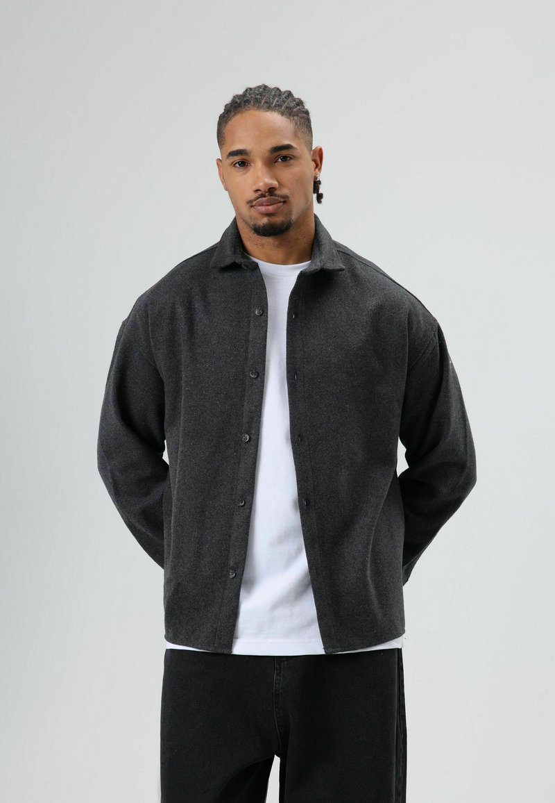 Grijs overhemd met knopen gemaakt van een zachte, gestructureerde stof met een omgeslagen kraag, gedragen over een wit T-shirt, gecombineerd met een zwarte broek.