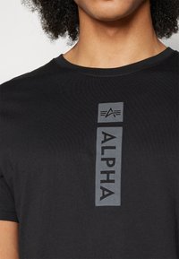 Mustan puuvillan t-paita, jossa on pystysuuntainen harmaa "ALPHA" -logo ja tyylitelty emblem. Pehmeä rakenne ja ympäri kaula-aukko. Normaalin mallin istuvuus.