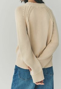 Person in beigem Strickpullover mit langen Ärmeln und blauen Jeans, steht mit verschränkten Händen hinter dem Rücken vor einem schlichten Hintergrund.