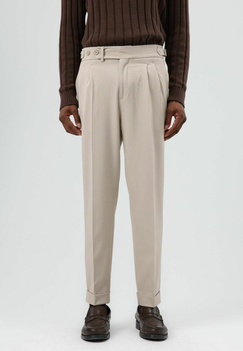 Beige op maat gemaakte broek met plooien, voorzien van een brede tailleband en knoopsluiting. Draagt een bruine geribbelde trui en donkere schoenen.