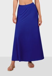 Dlhá, splývavá modrá sukňa vyrobená z hladkej látky, s širokým pásom a rozparkom, kombinovaná so zlatými sandálmi na remienky.