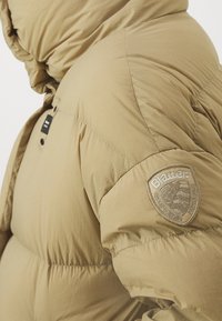 Khakifärgad vadderad jacka med huva, stora quiltade sektioner, tryckknappar och en axelbricka med varumärkeslogotyp. Mjukt och texturerat tyg.