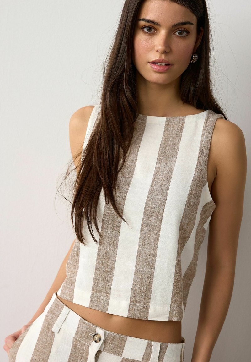 Noor naine, kellel on pikad tumedad juuksed, kannab varrukateta beeži ja valgeid vertikaalseid triibulisi üleriideid ja sobivaid pükse, seisab lihtsa hele tausta ees.