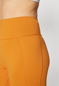 Høytlivde oransje leggings laget av elastisk stoff, med flate sømmer og en elastisk linning for komfort og passform.