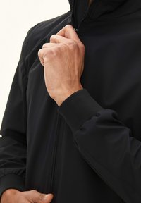 Sort zip-jakke med en glat overflade, elastiske manchetter og en afslappet pasform. Lynlåsen har et metallisk træk.