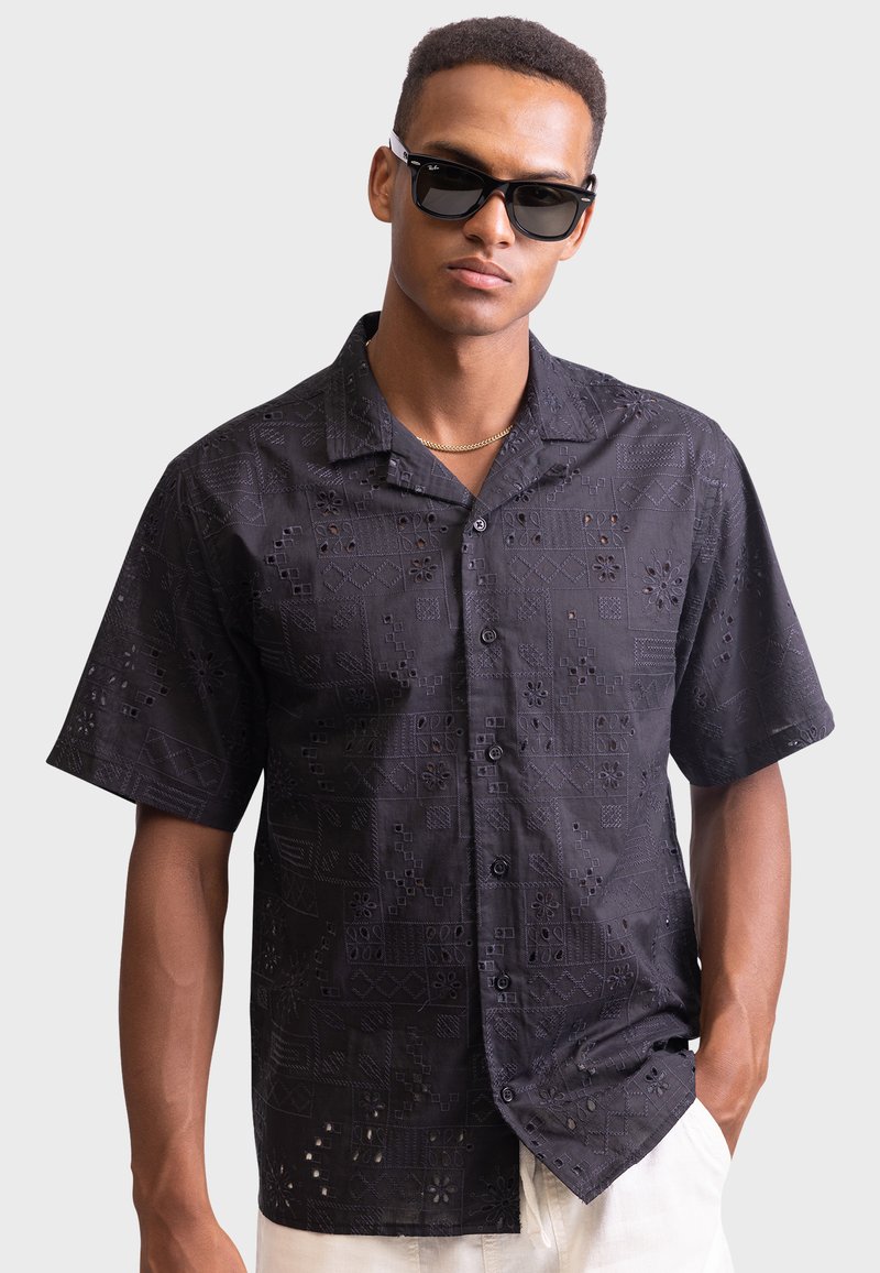Short-sleeve black shirt with cut-out patterns, button front, and collar. Model wearing black sunglasses with a neutral background.