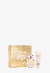 El conjunto de perfume Elie Saab incluye una botella de vidrio transparente con una tapa facetada, una caja dorada con intrincados patrones recortados y un tubo crema.