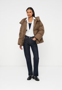 Brun pufferjakke med overdimensioneret hætte, hvidstribet skjorte, marineblå top og mørke denimjeans. Sorte sko fuldender outfittet.